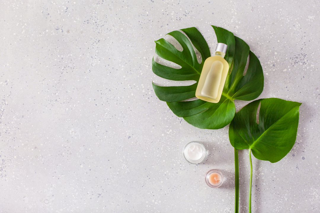 Bottle of skincare oil placed on large green tropical leaves, with two small jars on a light textured surface.