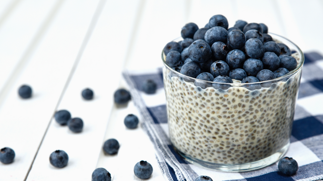 Glass jar of chia pudding topped with fresh blueberries, set on a striped cloth with scattered berries on a white wooden surface.