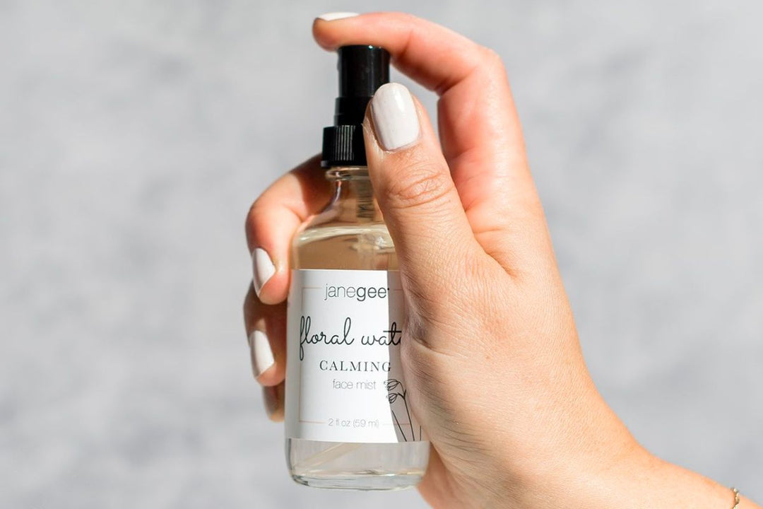 Hand holding a janegee calming floral water face mist bottle, pressing the spray pump against a light background.