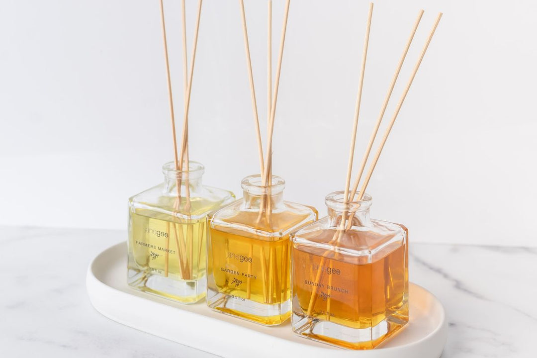 Three janegee reed diffusers in glass bottles with wooden sticks, displayed on a white tray against a clean background.