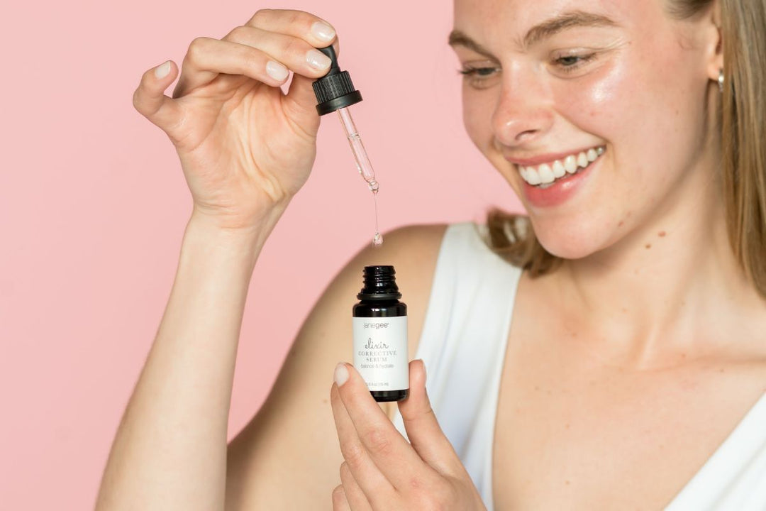 Woman smiles while holding a dropper above a janegee Elixir bottle, with liquid dropping against a soft pink background.