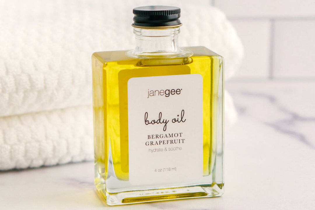 Glass bottle of janegee bergamot grapefruit body oil filled with golden liquid, set on a marble surface with a soft towel in the background.