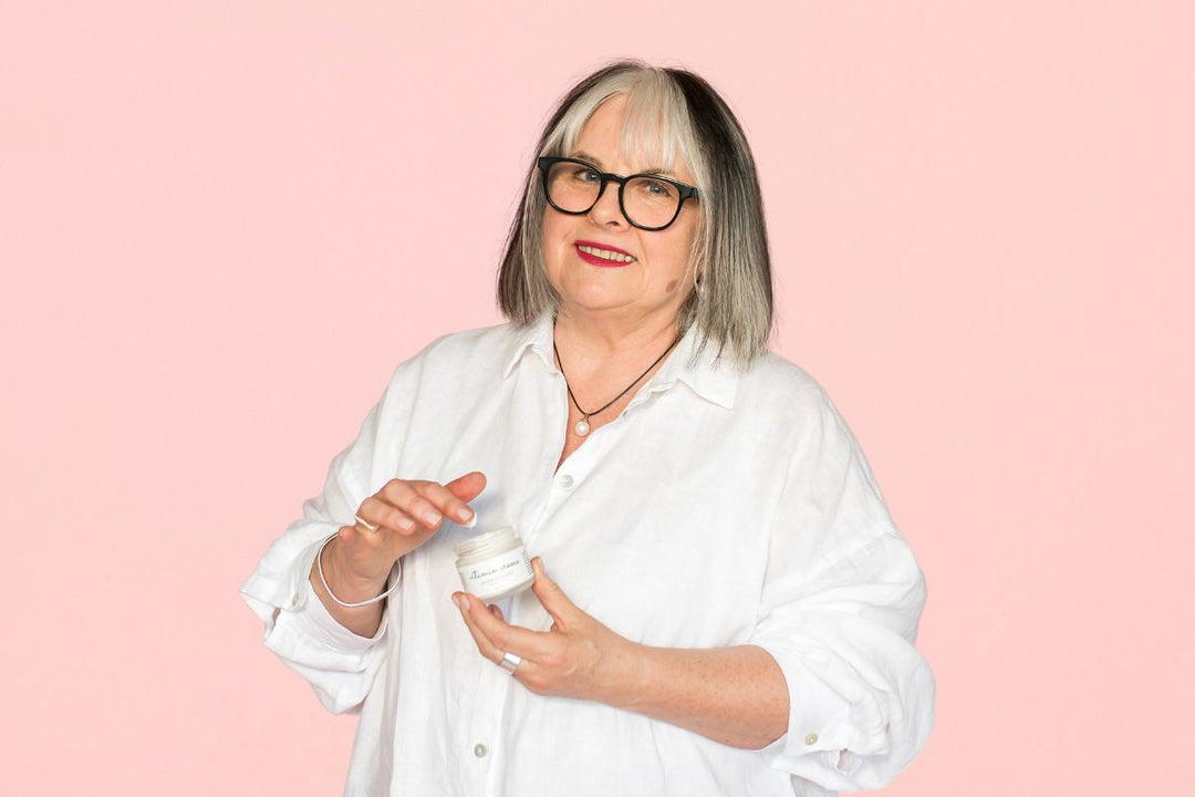 Woman in a white shirt holds a jar of vitamin cream, smiling against a soft pink background.
