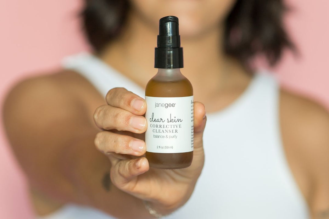 Hand holds a janegee clear skin corrective cleanser bottle against a soft pink background.