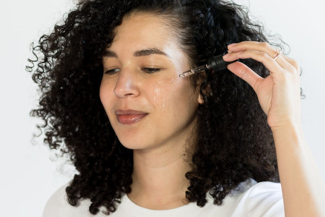 Woman applies Elixir with a dropper, serum visible on her cheek, highlighting a glowing skincare routine.