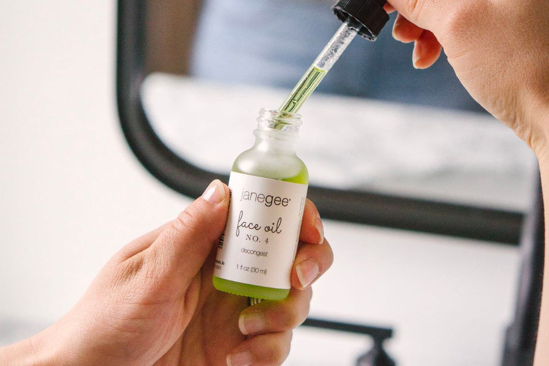Hand holding a janegee face oil No. 4 bottle while using a dropper to dispense green oil, with a softly blurred background.