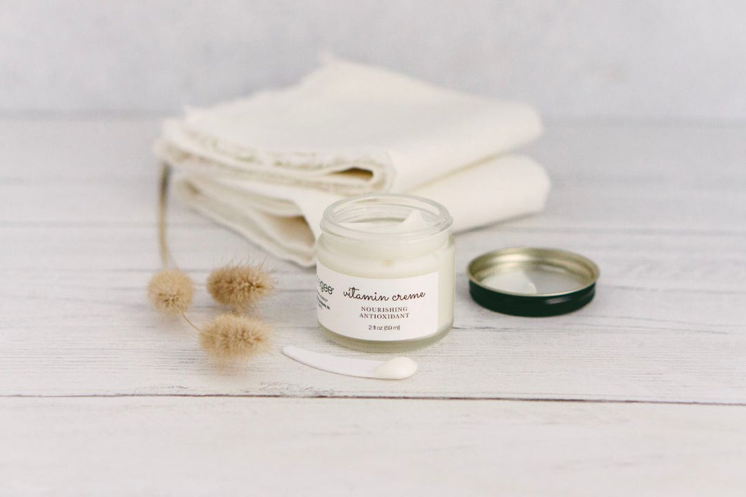 Open jar of janegee vitamin creme with a small dollop beside it, styled with folded cloths and dried botanicals on a light wood surface.