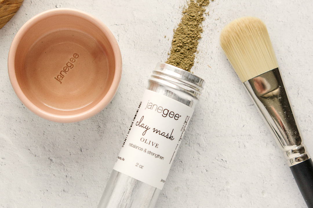 flat lay of a janegee olive clay mask in a metal bottle with loose powder spilled out, styled with a small ceramic mixing bowl and a facial brush on a neutral background