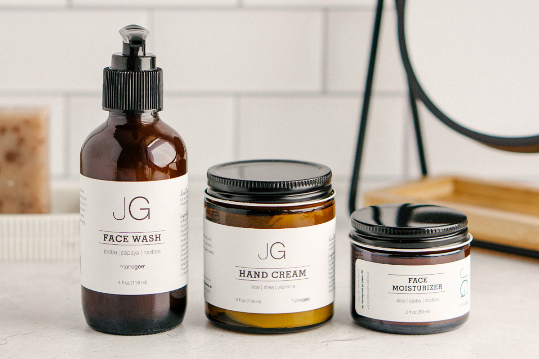 three amber glass skincare containers labeled JG including a face wash with pump, a hand cream jar, and a face moisturizer jar, styled on a bathroom counter with a clean, minimal background