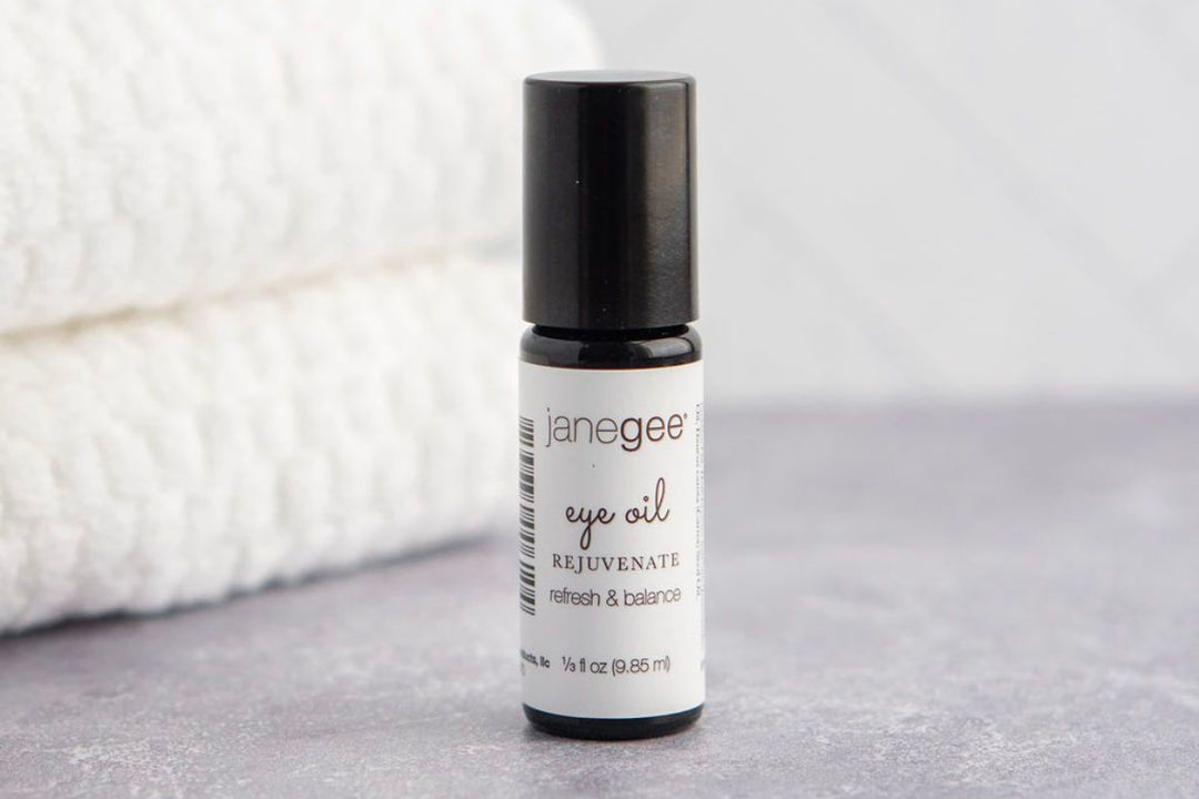 Small bottle of janegee rejuvenating eye oil with a black cap, placed on a neutral surface with a soft towel in the background.