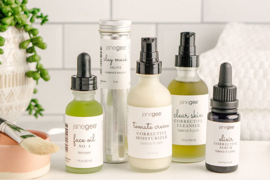 Collection of janegee skincare products including face oil, clay mask, moisturizer, cleanser, and serum arranged on a countertop with a brush and plant nearby.