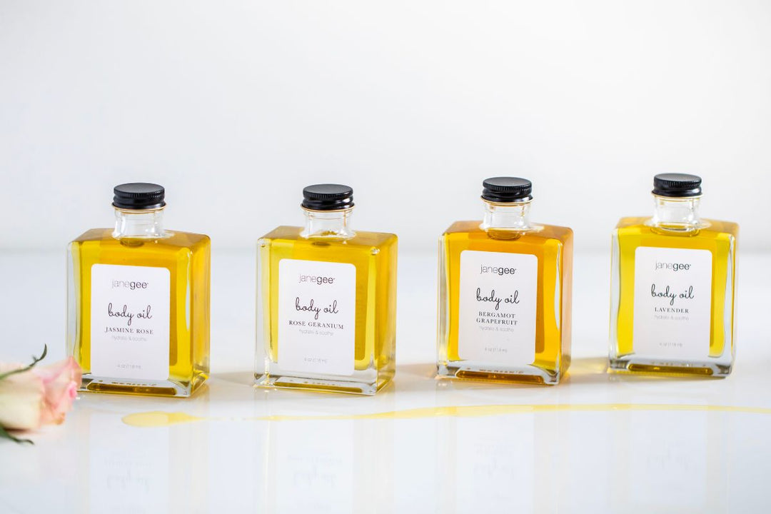 Four janegee body oil bottles in different scents arranged in a row on a white surface, with a small spill of oil and a flower nearby.