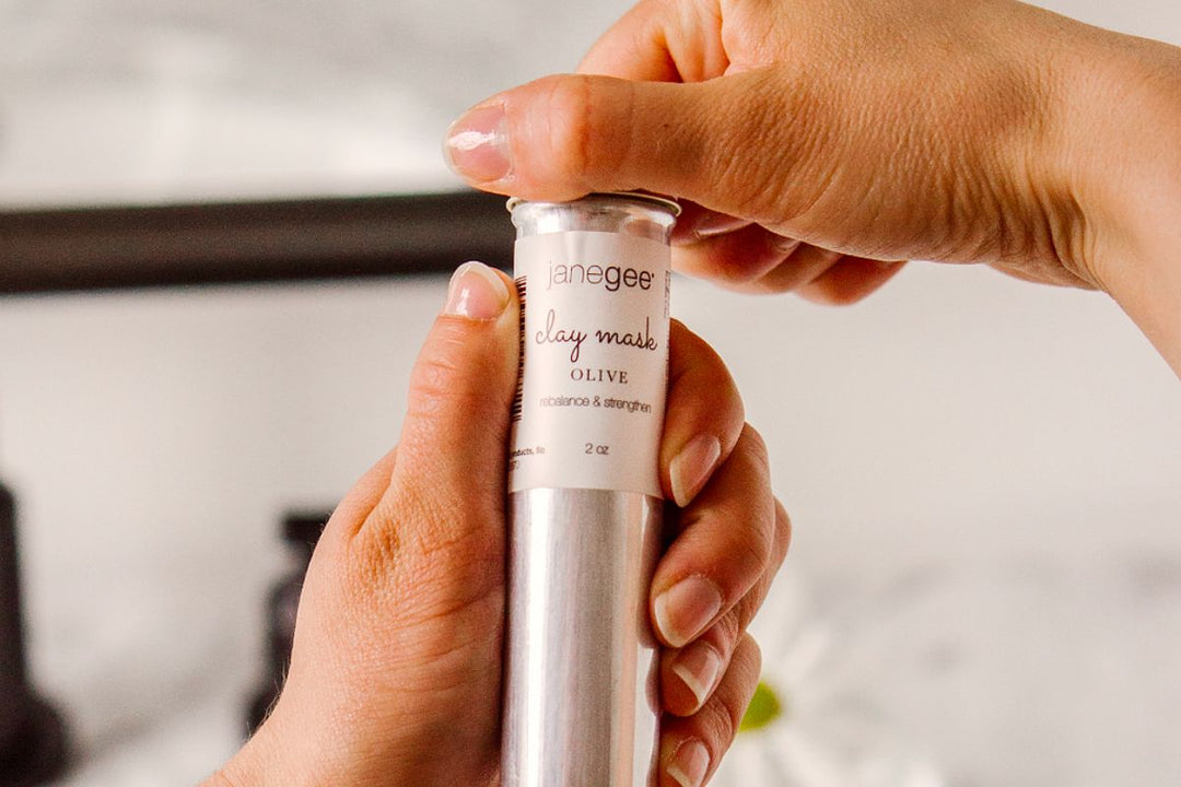 Hands opening a janegee olive clay mask tube, labeled “balance & strengthen,” against a softly blurred background.