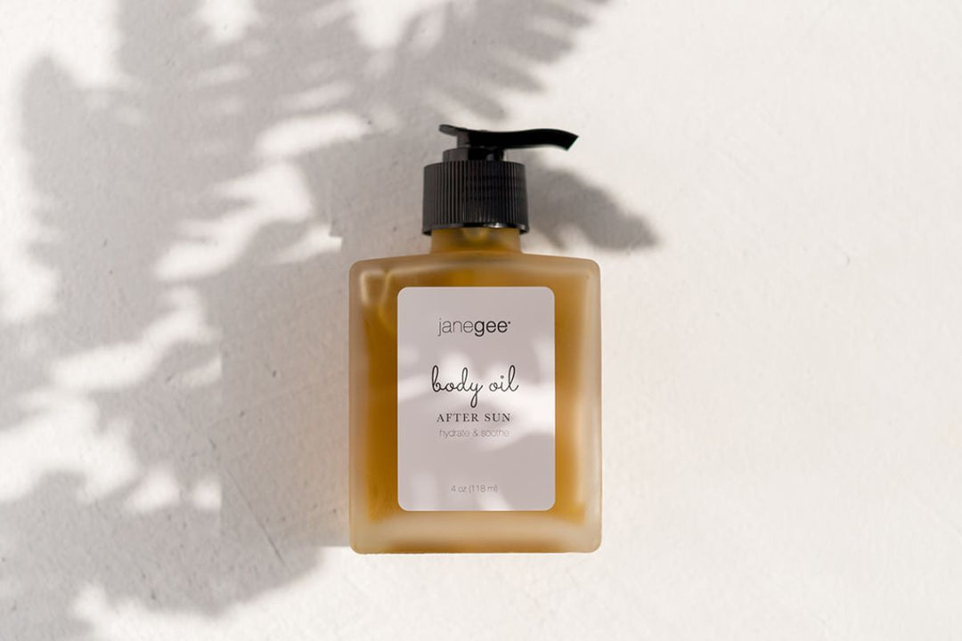 Bottle of janegee after sun body oil with pump top, casting soft leaf shadows on a light background.