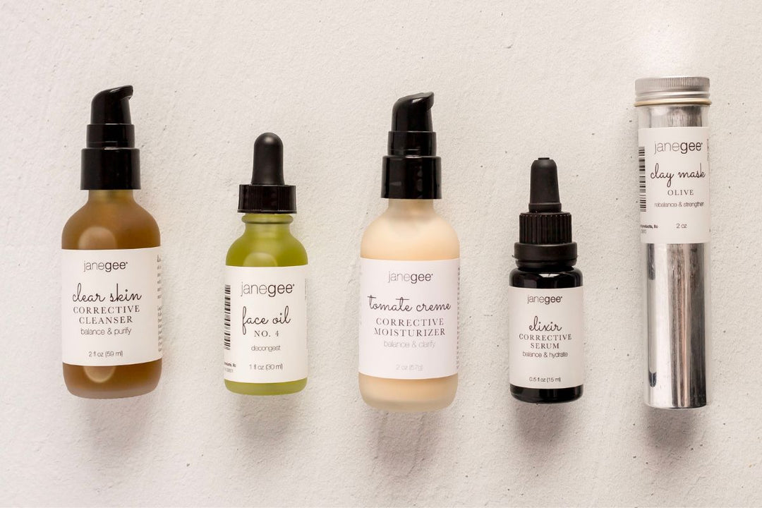 Lineup of janegee skincare products including clear skin cleanser, face oil No. 4, tomate creme moisturizer, elixir corrective serum, and olive clay mask on a neutral background.