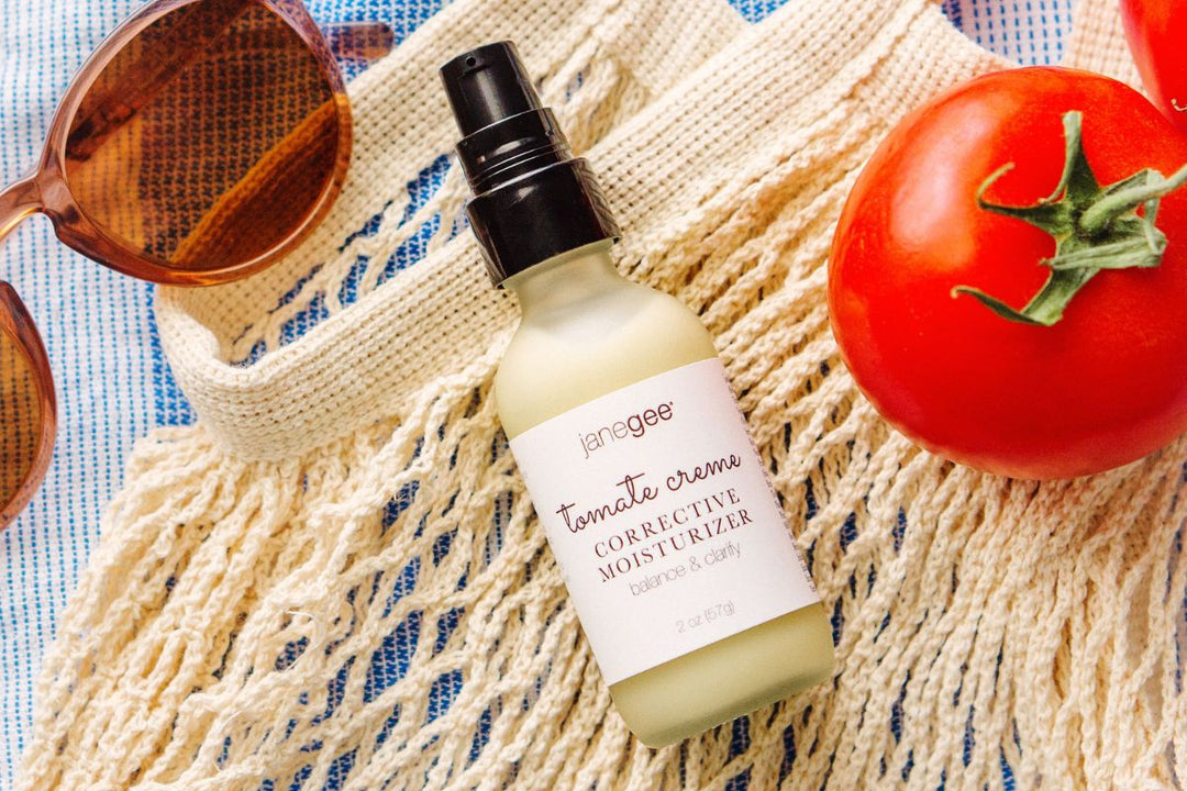 janegee tomate creme moisturizer bottle styled with a fresh tomato, sunglasses, and woven bag on a bright, summery background