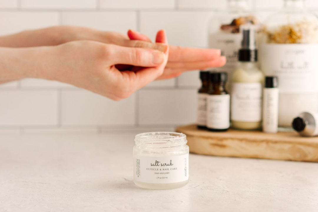 Hands rubbing product together above an open jar of janegee salt scrub for cuticle and nail care, with skincare bottles blurred in the background.