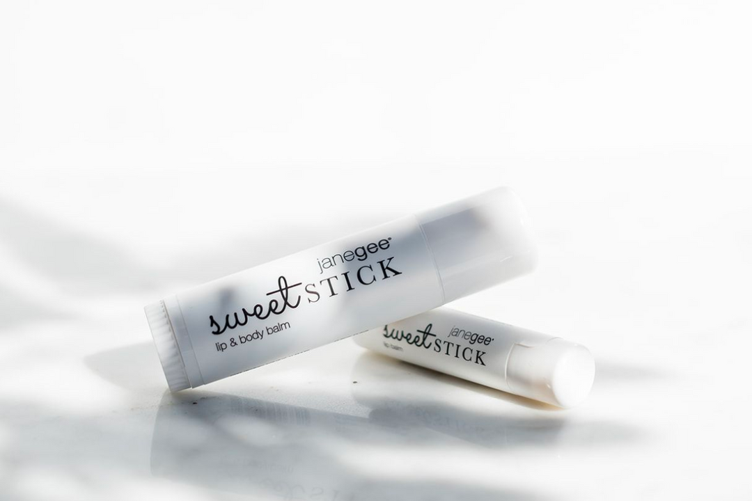 Two janegee Sweet Stick lip and body balm tubes on a white surface, softly lit with natural shadows.