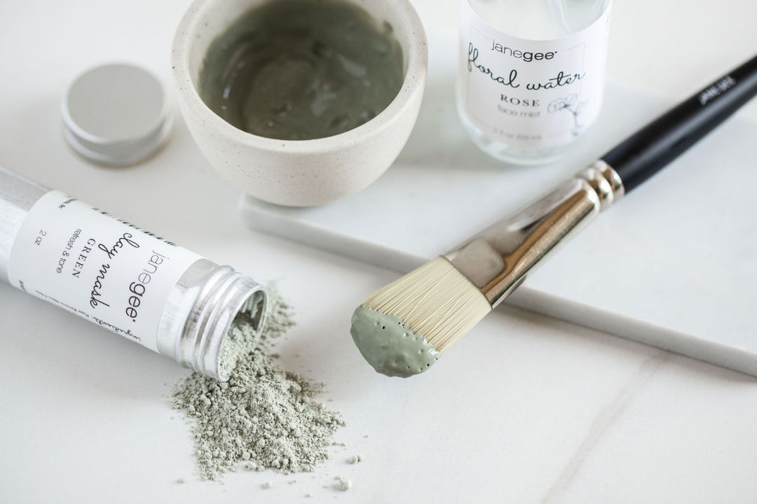 janegee green clay mask powder spilled beside a mixing bowl, brush with mask applied, and rose floral water on a clean surface