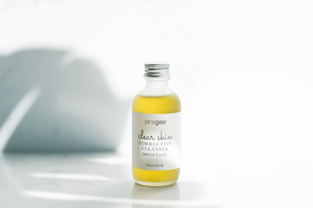 Small glass bottle of janegee clear skin corrective cleanser with metal cap, filled with golden liquid, set on a marble surface with soft natural light.