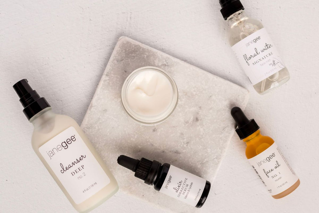 Flat lay of janegee skincare products including deep cleanser, face oil, elixir serum, and floral water arranged around an open jar of cream on a marble surface.
