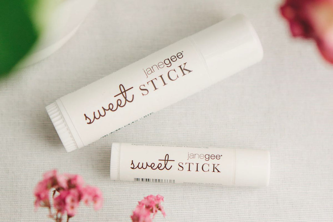two janegee sweet stick lip and body balms styled on a soft fabric background with delicate pink florals