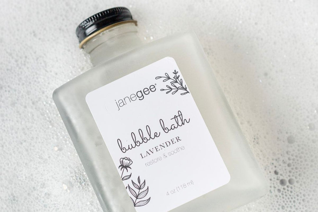 Frosted glass bottle of janegee lavender bubble bath resting in white bath foam, labeled “restore & soothe” with a black cap.
