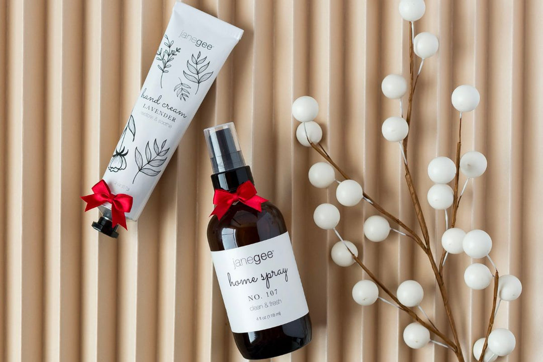 janegee lavender hand cream tube and home spray bottle styled with red ribbon bows on a neutral textured background with decorative white berries