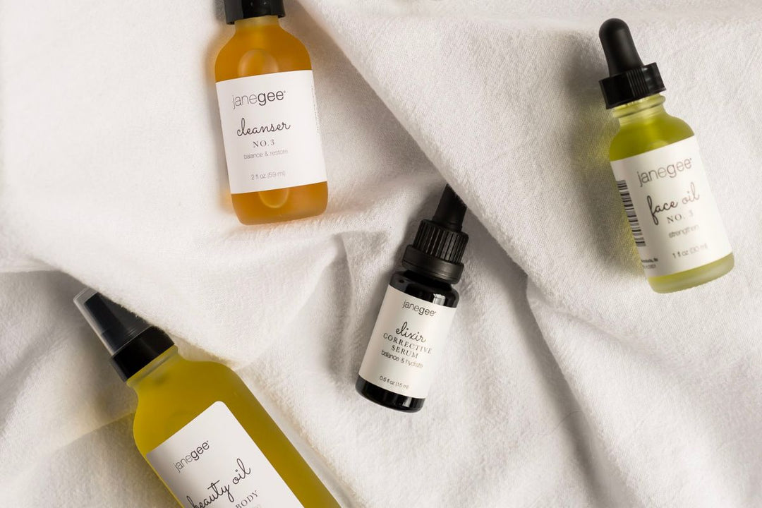 flat lay of janegee skincare essentials including cleanser, face oil, elixir corrective serum, and beauty oil arranged on soft white textured fabric