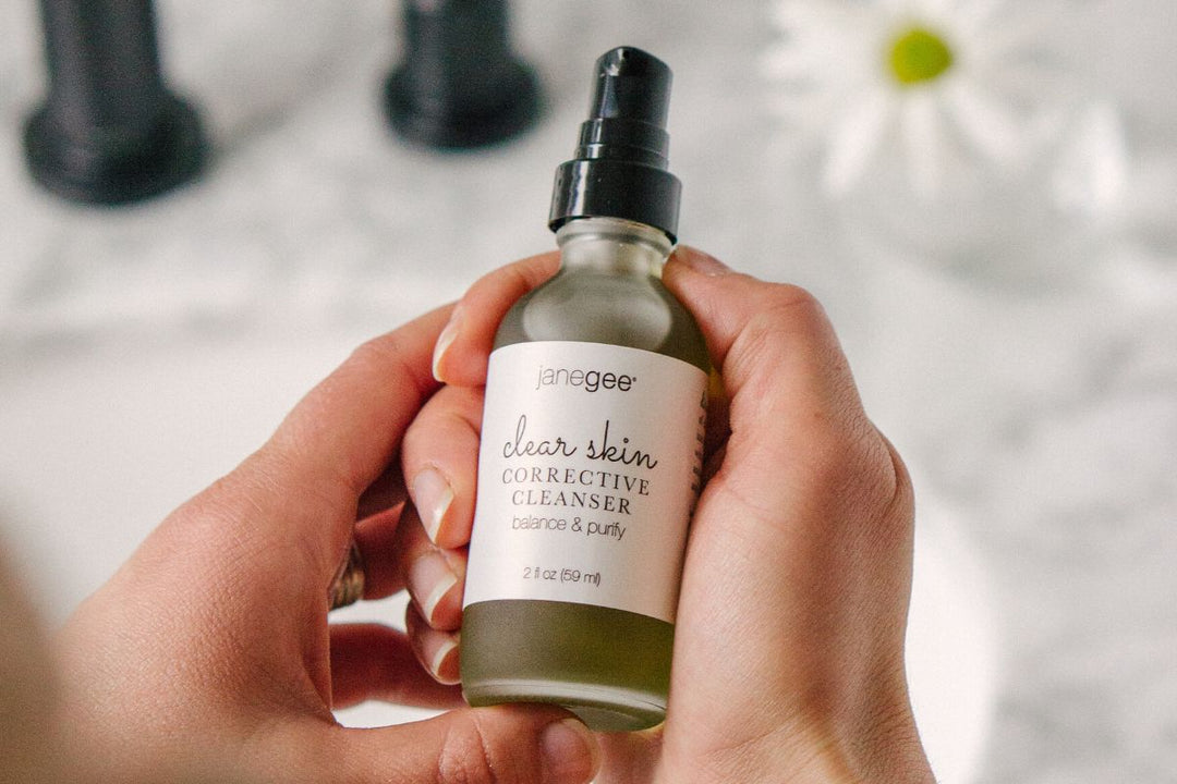 Hands holding a janegee clear skin corrective cleanser bottle with pump top, shown against a softly blurred background.