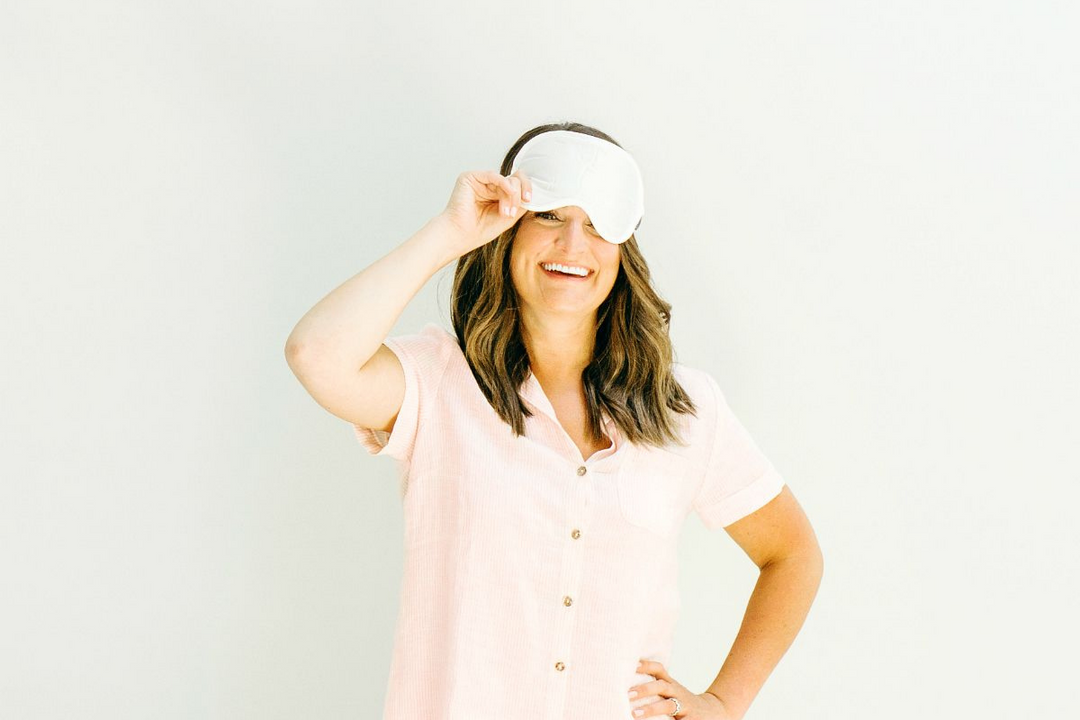 Smiling woman in a light pink shirt lifting a white sleep mask against a plain background.