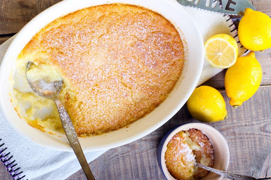 Baked lemon dessert in a dish dusted with powdered sugar, with a spoon scooping a serving and fresh lemons nearby on a wooden surface.