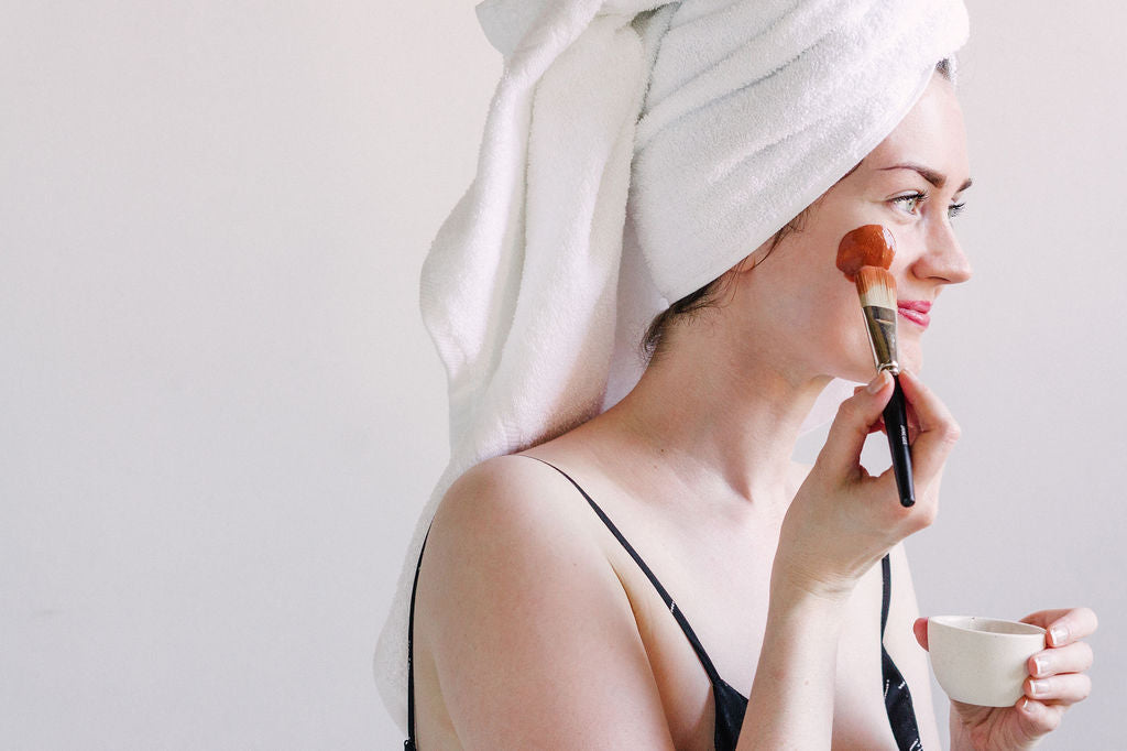 Woman with a towel wrapped around her hair applies a face mask with a brush while holding a small bowl, against a light background.