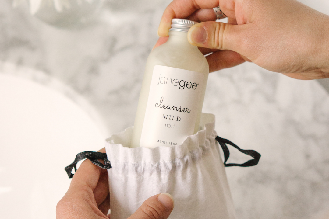 Hands placing a janegee Mild Cleanser bottle into a small drawstring pouch, set against a clean, minimal bathroom background