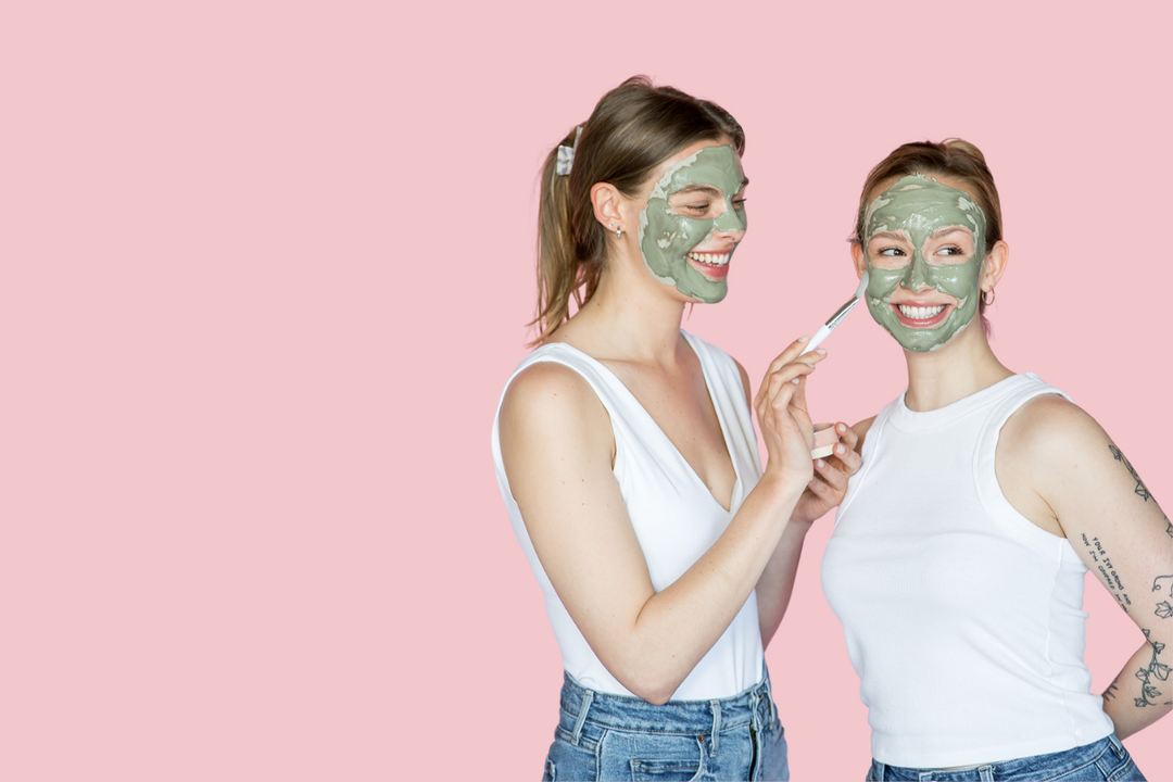 Two women in white tops apply green face masks to each other, smiling against a soft pink background.