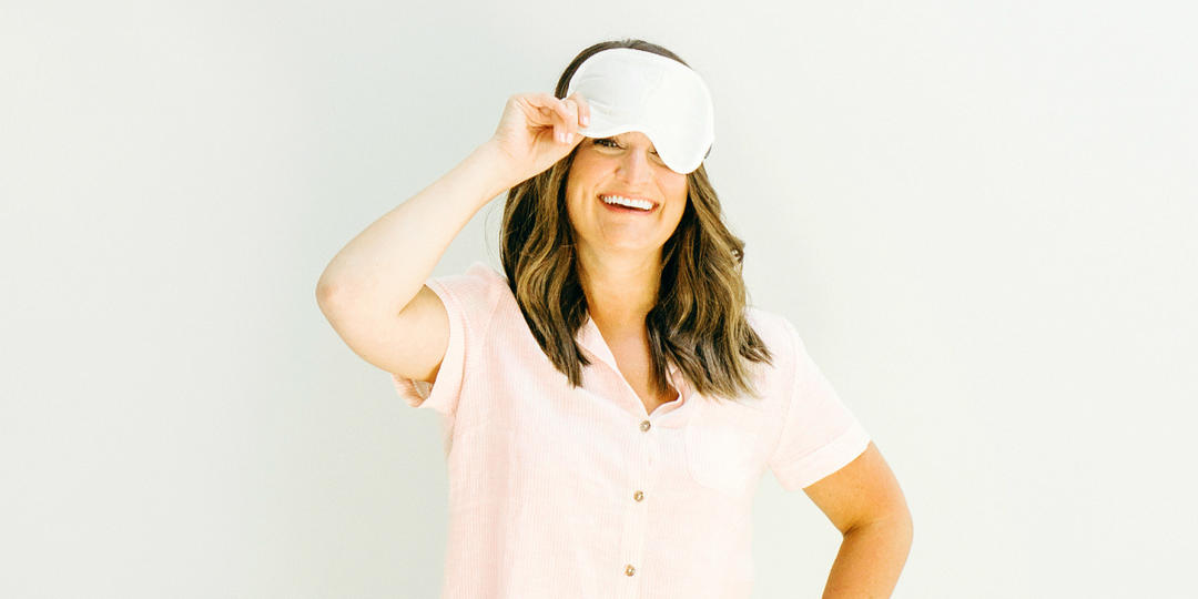 woman smiling and lifting a sleep mask, wearing a light pink top against a soft neutral background