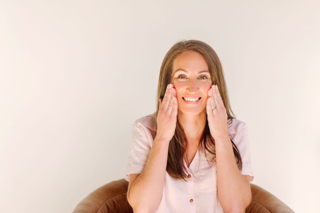Woman smiles while applying skincare to her cheeks, seated against a soft, neutral background.