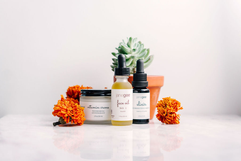 janegee skincare products including face oil, elixir, and vitamin creme arranged with marigold flowers and a potted succulent on a clean surface