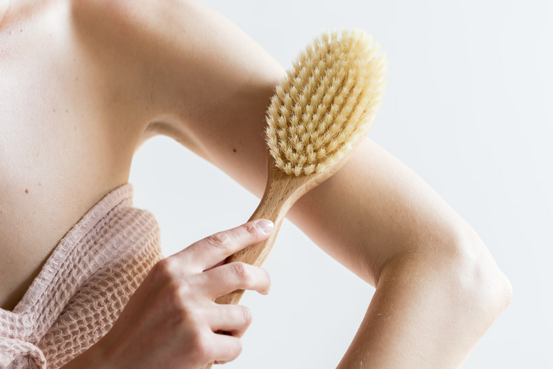 Person dry brushes their upper arm with a wooden brush, wrapped in a towel against a clean, neutral background.