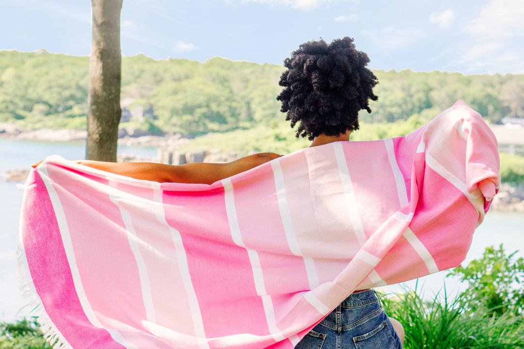 Person outdoors by the water holding a pink striped towel open across their back, with a scenic, sunlit landscape in the background.