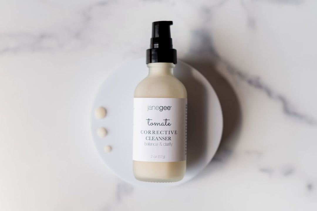 Bottle of janegee tomate corrective cleanser with pump top displayed on a white dish, with small drops of product beside it on a marble surface.