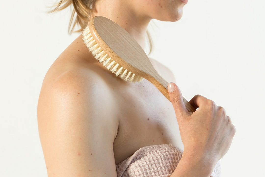 Person dry brushes their shoulder with a wooden brush, wrapped in a towel against a clean, neutral background.