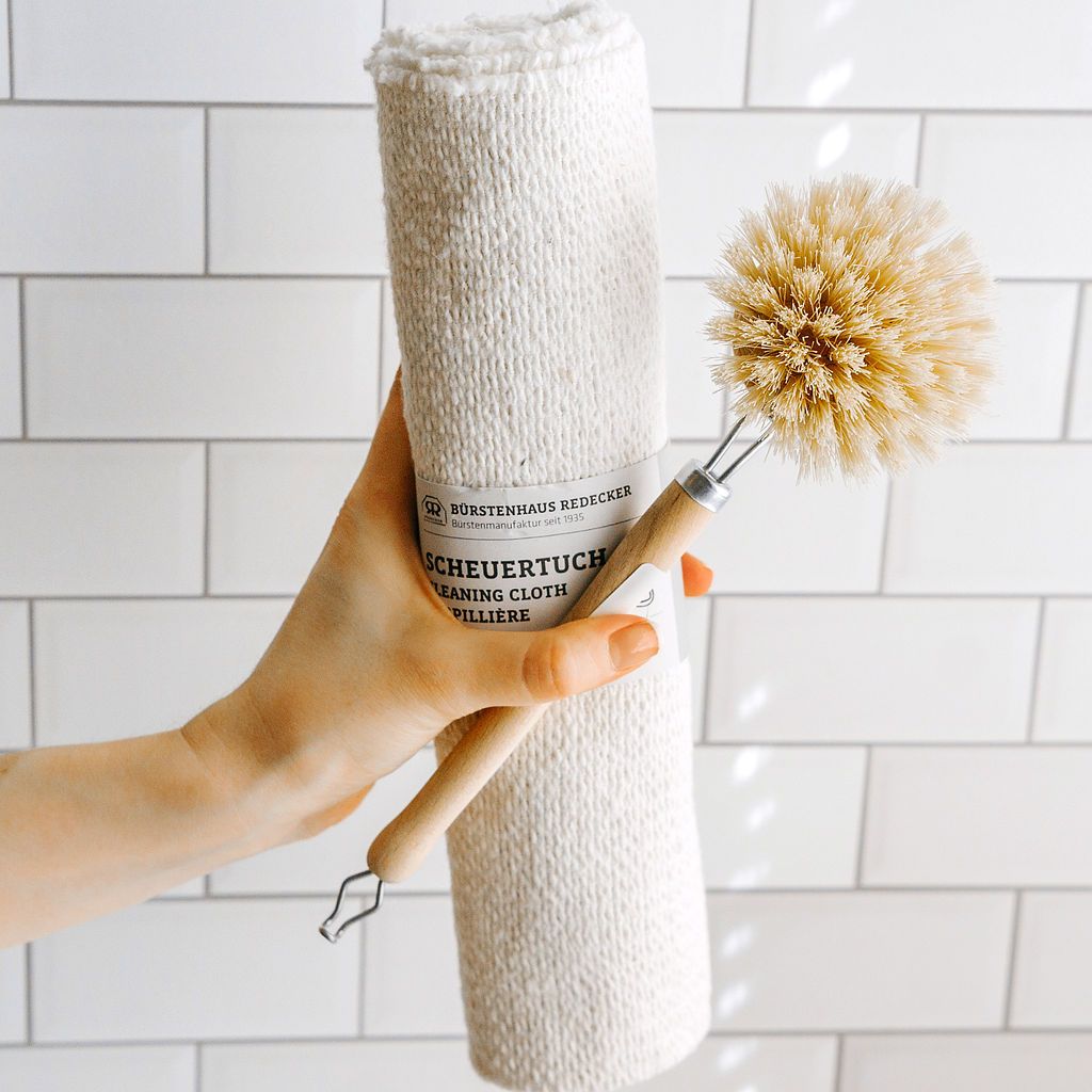 A hand holding a natural fiber cleaning brush and a rolled cotton cleaning cloth, showcasing simple, sustainable tools against a clean tiled background