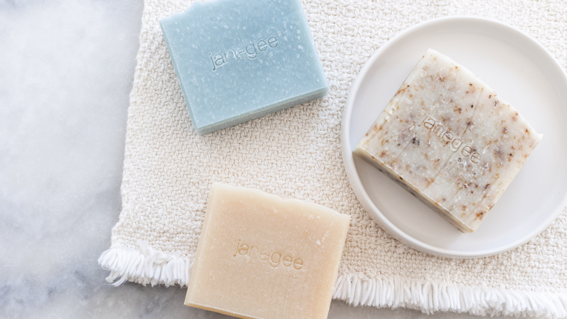 Three bars of soap on a textured white surface with 'janegee' branding.