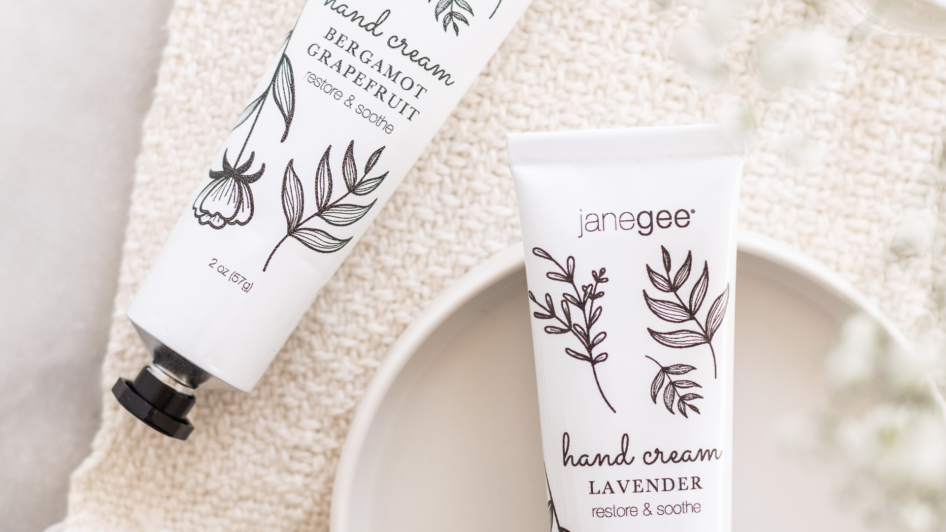 Two janegee bergamot grapefruit hand cream tubes with botanical illustrations on a textured cloth with one tube on a ceramic dish