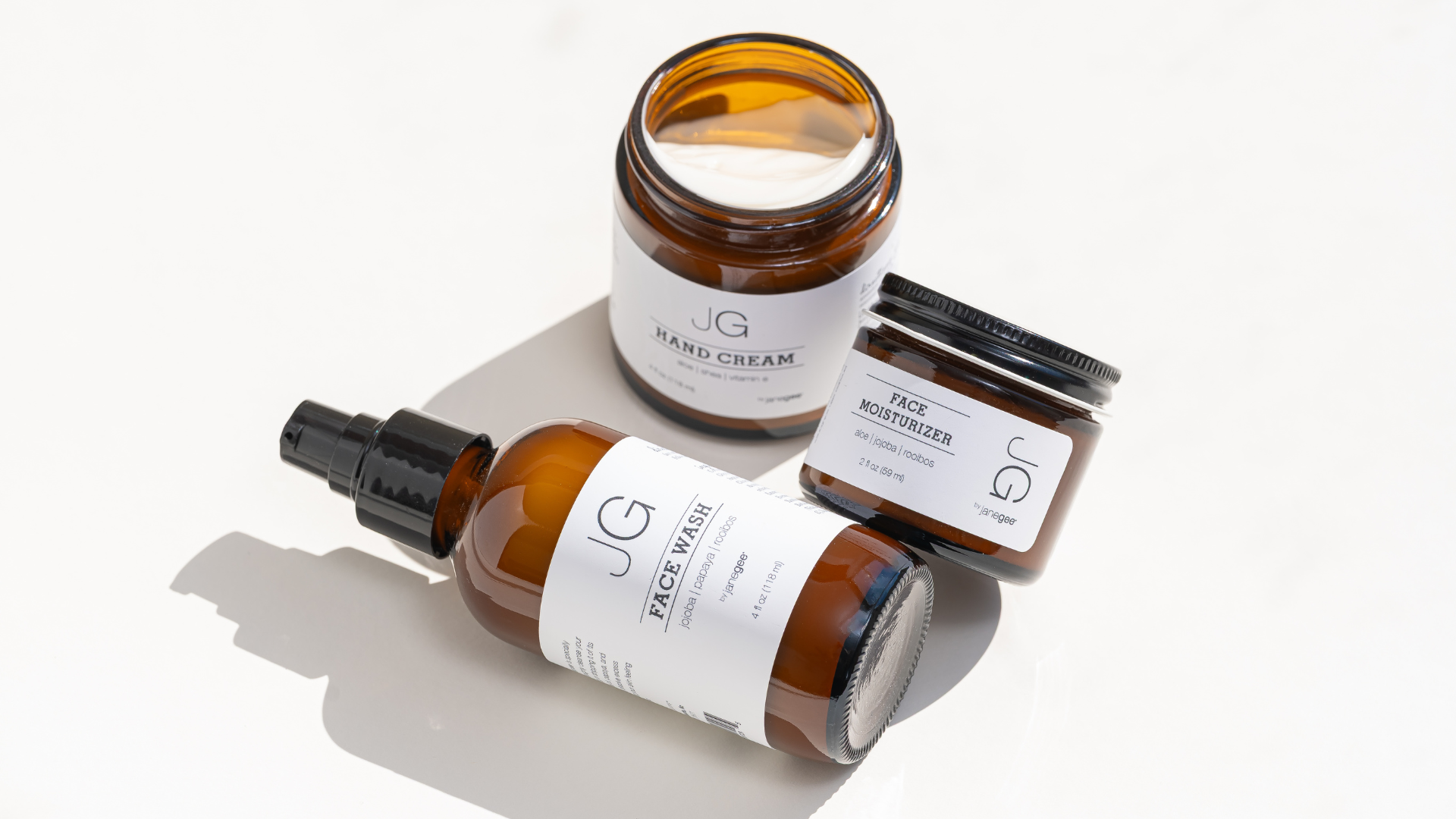 Amber glass skincare products arranged on a white surface, including an open jar of hand cream, a jar of face moisturizer, and a bottle of face wash. Soft natural light casts long shadows, highlighting the clean, minimalist packaging.