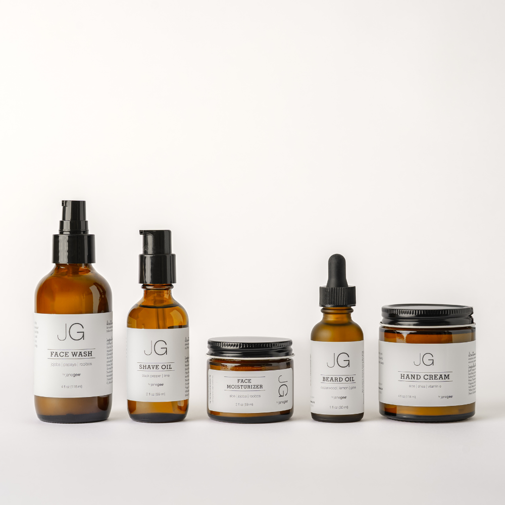 Collection of five janegee men's skincare products displayed on a white background, including Face Wash, Shave Oil, Face Moisturizer, Beard Oil, and Hand Cream in amber glass bottles and jars with minimalist labels.
