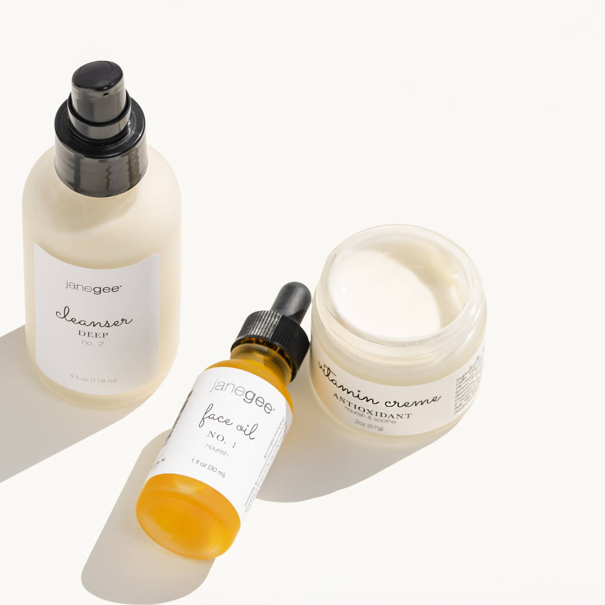 Three janegee skincare products arranged on a clean white surface in warm natural light: a bottle of Cleanser Deep No. 2, a bottle of Face Oil No. 1, and an open jar of Vitamin Crème Antioxidant moisturizer, highlighting their simple, minimalist packaging and nourishing skincare routine.
