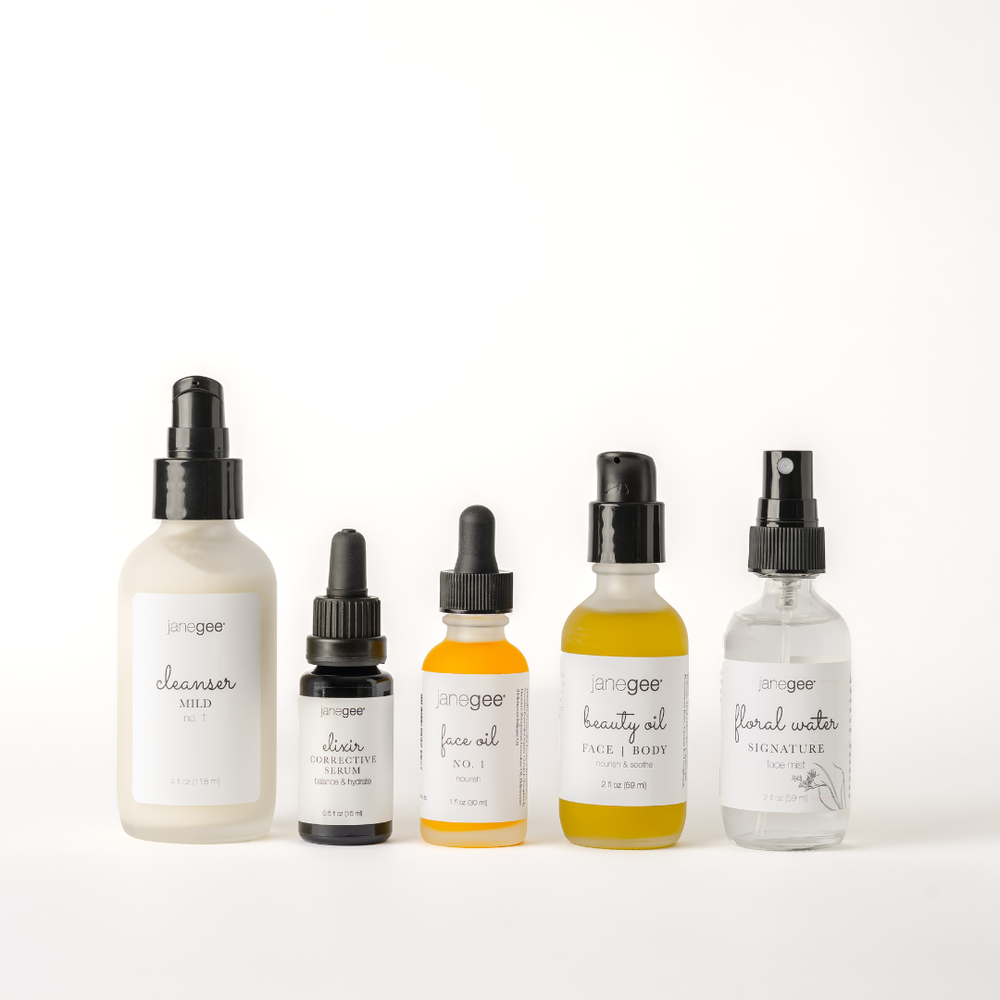 Five janegee skincare products displayed on a white background, including Cleanser Mild No. 1, Elixir Corrective Serum, Face Oil No. 1, Beauty Oil, and Signature Floral Water in frosted and clear bottles.