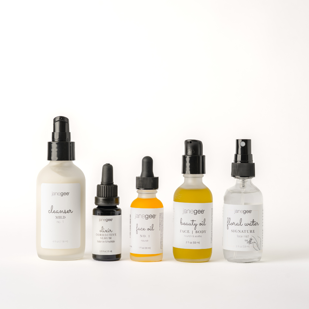Five janegee skincare products displayed on a white background, including Cleanser Mild No. 1, Elixir Corrective Serum, Face Oil No. 1, Beauty Oil, and Signature Floral Water in frosted and clear bottles.